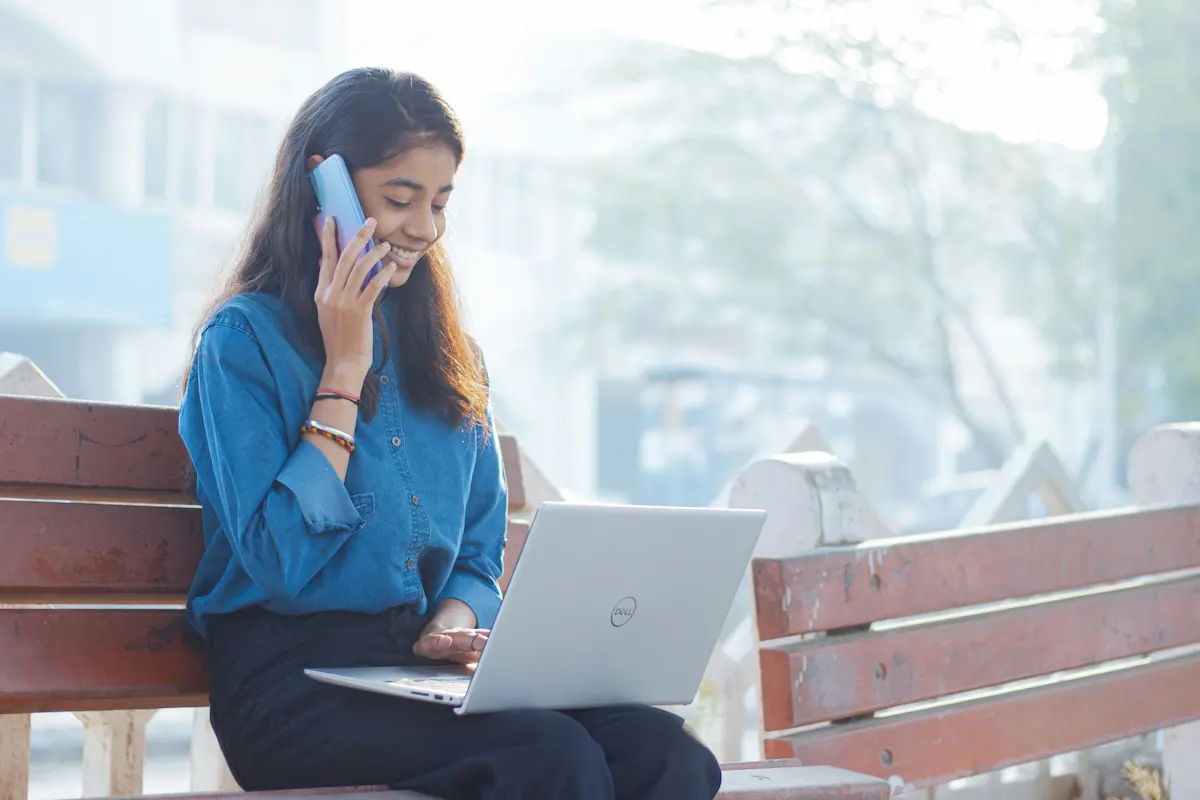 une femme assise sur un banc tenant son smartphone à l'oreille et travaillant sur son ordinateur portable